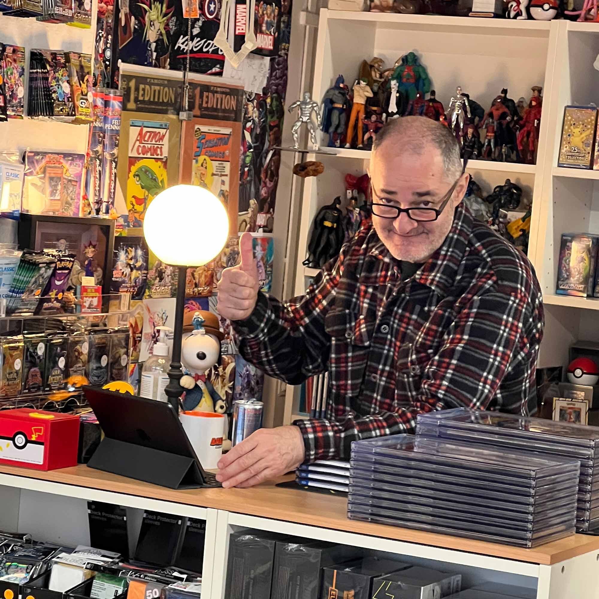 AMAZINGTOYS® Store Jahresrückblick 2024 Mann mit Brille zeigt hinter der Ladentheke im AMAZINGTOYS® Store den Daumen hoch, dahinter Actionfiguren